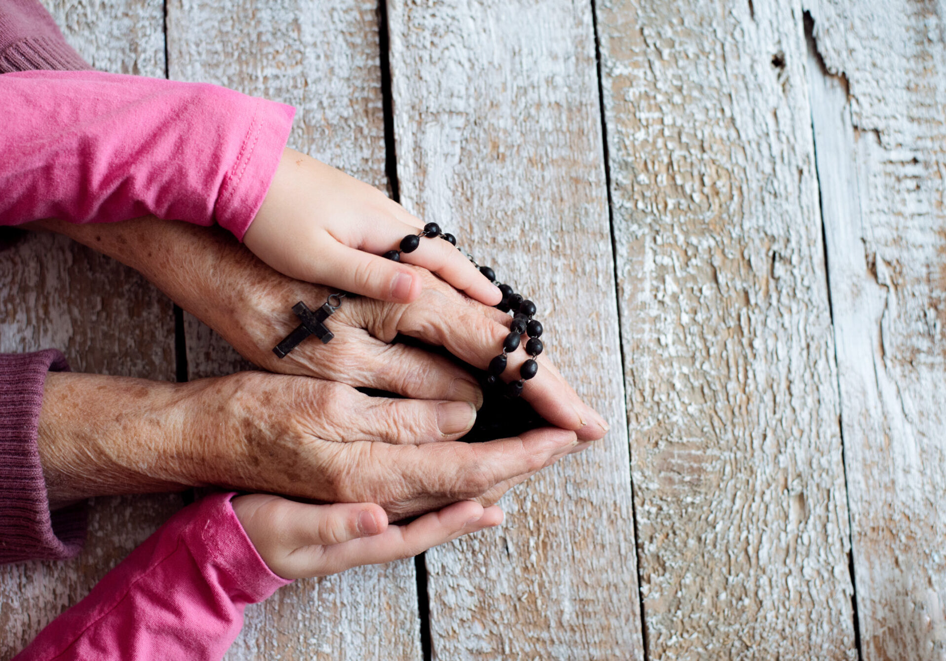 Close up. Hands of unrecognizable grandmother and her granddaughter holding rosary, praying. Studio shot on white wooden background.